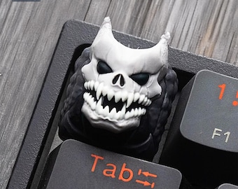 May include: A custom keycap featuring a white skull with sharp teeth and black eye sockets. The keycap is on a black keyboard, with the "Tab" key and "F1" key visible. The word "CAPSLOCK" is also visible.