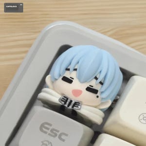 May include: A light blue anime-style character keycap with a white cape and a pink mouth. The keycap is on a white keyboard with the "Esc" key visible. The background is a light wood surface.