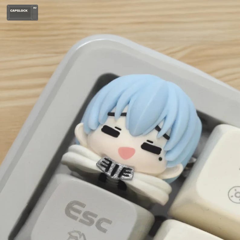 May include: A light blue anime-style character keycap with a white cape and a pink mouth. The keycap is on a white keyboard with the "Esc" key visible. The background is a light wood surface.