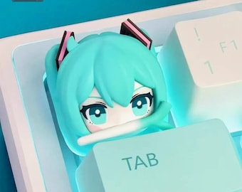 May include: A custom keyboard keycap featuring a turquoise anime character's face with pink accents. The keycap is placed on a white keyboard, with the "TAB" key visible. The background is a light blue.