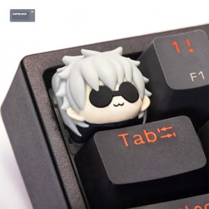 May include: A custom keycap for a mechanical keyboard, depicting a cartoon character with white hair and black sunglasses. The keycap is set in a black keyboard, with the orange "Tab" key visible. A unique keyboard accessory.