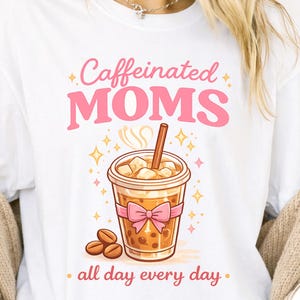 May include: White t-shirt with a graphic of an iced coffee drink, coffee beans, and the text "Caffeinated MOMS all day every day" in pink. The iced coffee has ice cubes and a straw, with a pink bow around the cup.