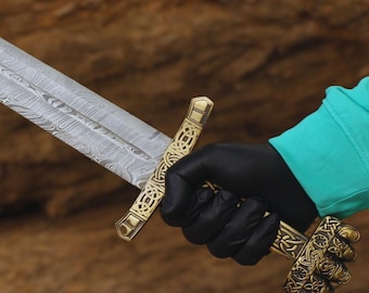 May include: A long, silver sword with a detailed blade pattern and a gold-coloured hilt. The sword is held by a gloved hand against a wooden background. The hilt features intricate designs.