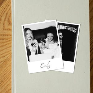 Puede incluir: Dos fotos en blanco y negro estilo Polaroid sobre un libro verde claro. Las fotos muestran a personas sosteniendo copas de vino. Una foto tiene el nombre "Emily" escrito. El libro está sobre una superficie de madera.