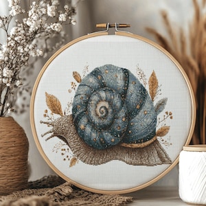 May include: A cross-stitch embroidery hoop with a detailed snail design. The snail has a blue shell with gold accents, surrounded by brown leaves and floral elements. The embroidery is set in a wooden hoop, creating a rustic, handmade aesthetic.