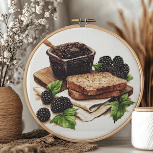 May include: A circular embroidery hoop containing a cross-stitch pattern of a square jar filled with dark blackberry jam, two slices of toasted bread, and several fresh blackberries with green leaves, all set against a white fabric background.
