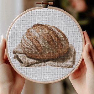 May include: A cross-stitch embroidery of a loaf of bread on a linen cloth, displayed in a wooden embroidery hoop. The bread is brown with lighter highlights, and the cloth is a neutral tan color.