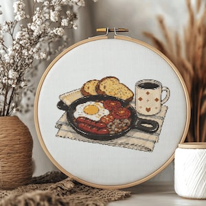 May include: A circular wooden embroidery hoop containing a cross-stitch design of a breakfast scene. The image features a black skillet with a fried egg, sausages, and tomatoes, alongside two slices of toasted bread and a patterned coffee mug.