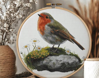 Pode incluir: Bordado em ponto cruz de um pássaro robin numa rocha com margaridas, num bastidor de madeira. O pássaro tem peito vermelho, asas verdes e penas cinzentas. O bordado evoca um tema rústico, inspirado na natureza.