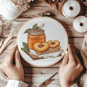 May include: An embroidery hoop containing a fabric panel with a cross-stitch design of a glass jar filled with orange peach jam, a silver spoon, and two peach halves resting on a slice of bread, all set against a white background.