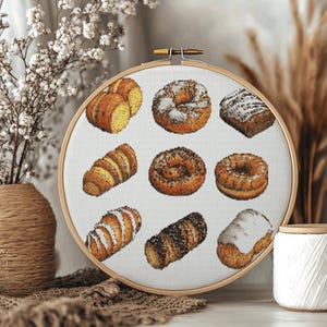 Puede incluir: Un bastidor de bordado de punto de cruz con varios productos horneados, incluyendo cruasanes, donuts y pasteles. El bordado está sobre un fondo blanco, dentro de un aro de madera. La paleta de colores incluye tonos marrones, beige y blanco.