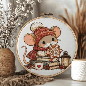 May include: A circular wooden embroidery hoop containing a cross-stitch pattern of a mouse wearing a red knit hat and scarf. The mouse sits on books, holding a snowflake-shaped cookie next to a small mug with a red heart and a lit lantern.
