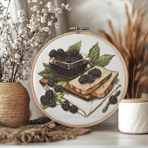 Op de afbeelding: Een ronde houten borduurring met een kruissteekpatroon van een glazen kom gevuld met donkere bramen, twee sneetjes brood belegd met bramen en losse bessen op een wit doek, alles afgebeeld op een witte stoffen achtergrond.