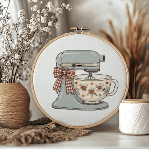 May include: A circular wooden embroidery hoop containing a cross-stitch pattern of a light blue stand mixer. The mixer features a red and white gingham bow on its side and holds a white mixing bowl decorated with a red and green floral pattern.