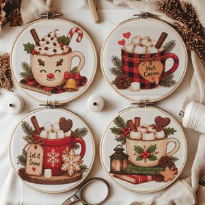 May include: Four circular embroidery hoops featuring holiday-themed designs of mugs filled with hot cocoa, marshmallows, and festive decorations. The designs include text reading "Hot Cocoa", "Let it Snow", and "Warm & Cozy" with winter motifs.
