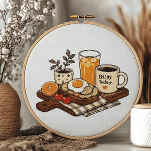Puede incluir: Un bastidor de bordado circular de madera que contiene un patrón de punto de cruz de una escena de desayuno sobre tela blanca. El diseño presenta una tabla de cortar de madera con un huevo frito sobre tostada, un vaso de zumo de naranja, una pequeña planta y una taza con el texto "ENJOY today" y un pequeño corazón naranja.