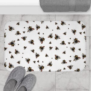 May include: White rectangular bath mat with a repeating pattern of bumblebees in various sizes. The bees are black and yellow with brown wings. The mat is on a tiled floor, with gray slippers in the foreground.
