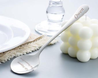 May include: A silver spoon engraved with the name "Owen" on the handle. The spoon's bowl has the text "Life Happens Eat Dessert First". A white bubble candle and a white plate are in the background.
