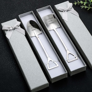 May include: Four silver-colored shovel-shaped spoons, each in a gray gift box with a silver bow. The spoons have the text "CUSTOM TEXT HERE" engraved on the shovel head. The boxes are arranged on a black surface.