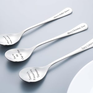 Może przedstawiać: Trzy łyżki ze stali nierdzewnej z wygrawerowanym tekstem. Na jednej łyżce widnieje napis "Mom's Coffee Spoon", na drugiej "Merry Christmas, Love", a na trzeciej "Zero Calorie Zone". Łyżki leżą na jasnoszarej powierzchni.