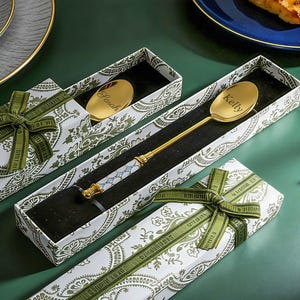 Puede incluir: Cucharas y palillos dorados presentados en cajas decorativas con cinta verde. La cuchara tiene grabado el nombre "Kelly". Las cajas tienen un estampado de cachemira blanco y verde.