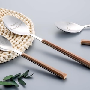 Op de afbeelding: Roestvrijstalen lepels met bruine houten handvatten liggen op een lichtgrijze ondergrond. De lepels hebben gegraveerde teksten: "Made with Love & Caffeine" en "Custom Spoon, Any Text You Want". Een geweven placemat en groene bladeren zijn ook zichtbaar.