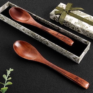 Op de afbeelding: Twee houten lepels met gegraveerde tekst, één met "Sweet Kitchen" en de andere met "Martin". De lepels worden gepresenteerd in een decoratieve doos met een paisley patroon en een groen lint. De lepels hebben een warme bruine kleur.