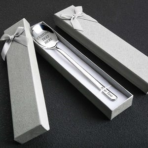 Może przedstawiać: Srebrzysta metalowa łyżka z wygrawerowanym napisem CUSTOM TEXT HERE na czerpaku oraz Your Text na trzonku, umieszczona w długim, jasnoszarym, fakturowanym pudełku prezentowym z pasującą kokardą na wieczku.