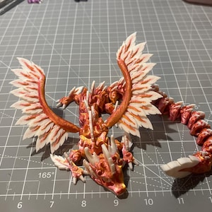 May include: A multi-jointed dragon figurine featuring a vibrant orange and yellow gradient body with white accents on the wings, horns, and tail tip. The creature has large, feathered wings extended upward and a long, segmented body that curves along the surface.