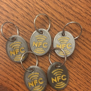 May include: Five oval-shaped gray keychains with silver metal rings. Each keychain features a yellow embossed NFC logo with concentric signal waves centered on the surface. The texture of the gray material shows visible 3D printed layer lines.