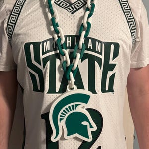 Puede incluir: Camiseta de malla blanca con "MICHIGAN STATE" y el número "12" en verde. Se lleva un collar de cadena grande verde y blanco sobre la camiseta. El collar tiene un colgante con el logotipo de un casco espartano.