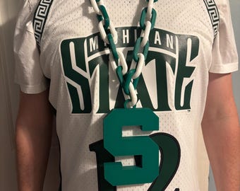 May include: A white mesh jersey with green lettering that says "MICHIGAN STATE" and the number "12". A large green and white chain necklace with a green "S" pendant is worn over the jersey.