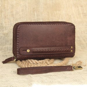 May include: A dark brown leather wallet with a zip closure and a wrist strap. The wallet has a textured surface and is accented with brass-coloured rivets and stitching. A braided leather detail adds a decorative touch.