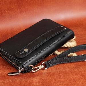 May include: Black leather wallet with a wrist strap. The wallet has a zipper closure and decorative stitching. The wrist strap is attached with a silver-colored clasp. The wallet is on a brown surface.