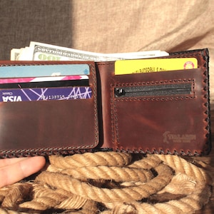 May include: Open brown leather wallet with multiple card slots, a zippered pocket, and cash. The wallet is hand-stitched with a dark brown thread and has a textured appearance. The wallet is open, displaying its contents.