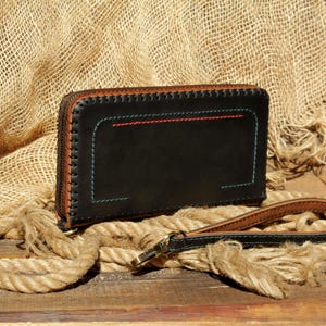 May include: Black leather wallet with a brown zipper and accents. The wallet features red and teal stitching details. A matching wrist strap with a metal clasp is also visible. The wallet is displayed on a wooden surface with rope.
