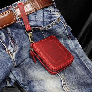Op de afbeelding: Een rode leren portemonnee met ritssluiting en bijpassende polsband is bevestigd aan een riemlus. De portemonnee hangt aan een bruine leren riem, tegen een achtergrond van blauwe spijkerbroek.