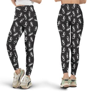 Puede incluir: Leggings negros con un estampado repetido de lagartos blancos. Los leggings de cintura alta se muestran desde la parte delantera y trasera, destacando el estampado integral. Los leggings se usan con zapatillas.