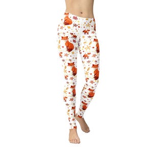 May include: White leggings with a repeating pattern of orange foxes, autumn leaves, and star shapes. The leggings are made of a stretchy material and are designed to fit snugly. The overall theme is fall and nature.