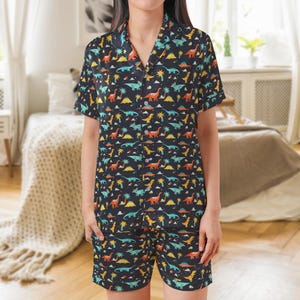 May include: Black pajama set with a colorful dinosaur pattern. The short-sleeved top and shorts feature a repeating design of various dinosaurs, palm trees, and stars in shades of orange, yellow, blue, and green.