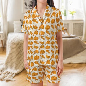 May include: A two-piece pajama set featuring a white background with a repeating pattern of fried chicken pieces in shades of brown and yellow. The set includes a button-down shirt with short sleeves and matching shorts.