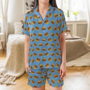 May include: Light blue pajama set featuring a repeating pattern of cartoon hamburgers. The set includes a short-sleeved button-down shirt and matching shorts. The pajamas have a classic collar and are made of a soft, lightweight fabric.