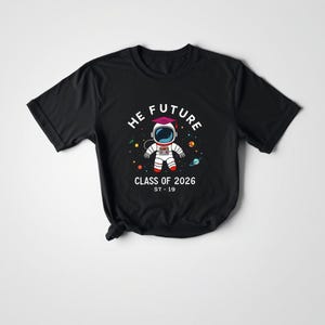 Peut inclure: T-shirt noir avec un astronaute de dessin animé et le texte "HE FUTURE" et "CLASS OF 2026". L'astronaute est entouré de planètes et d'étoiles colorées. Le t-shirt porte également le texte "ST-19".
