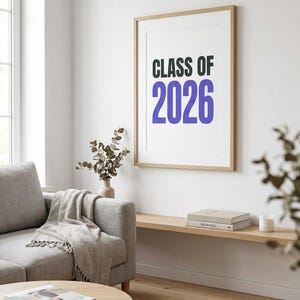 Peut inclure: Une affiche encadrée avec le texte "CLASS OF 2026" en noir et bleu. L'affiche est accrochée à un mur blanc au-dessus d'une étagère en bois avec des livres et des bougies. Un canapé gris, une table basse et un tapis sont au premier plan.