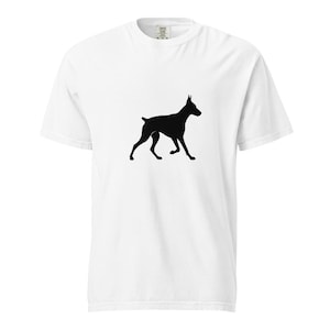 Può includere: T-shirt bianca con una silhouette nera di un cane Dobermann Pinscher. Il cane è raffigurato in posizione di camminata, con le orecchie dritte e la coda leggermente sollevata. La maglietta è realizzata in un materiale morbido e confortevole.