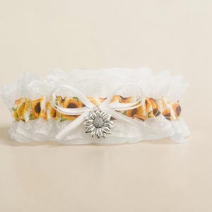 May include: A white lace garter featuring a ruffled trim and a central band patterned with yellow and brown sunflowers. A white satin ribbon is tied into a bow at the center, accented with a silver-toned metal sunflower charm hanging from the knot.