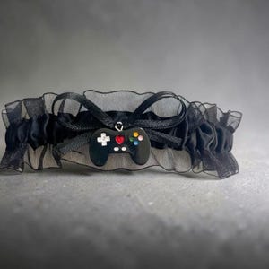 May include: A black garter featuring a ruffled sheer fabric band and a central black satin ribbon bow. A small, glossy black game controller charm hangs from the center, detailed with a white directional pad, a red heart, and four colorful buttons.