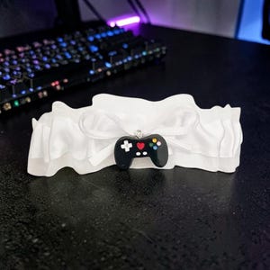 May include: A white ruffled fabric garter featuring a central white ribbon bow. Attached to the bow is a black video game controller charm with a red heart in the center, a white directional pad, and small colorful circular buttons on the right side.