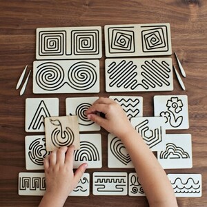 May include: A collection of light-colored wooden tracing boards featuring black engraved patterns including spirals, geometric shapes, waves, and floral designs, accompanied by two small wooden styluses for tactile practice and pattern tracing.