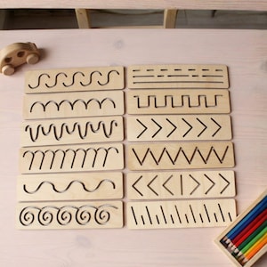 Könnte beinhalten: Ein Set aus zwölf rechteckigen, hellen Holztafeln zum Nachspuren mit verschiedenen eingravierten Mustern wie Wellen, Zickzacklinien, geraden Linien und geometrischen Formen, ergänzt durch ein kleines Holzauto und eine Schachtel Buntstifte.